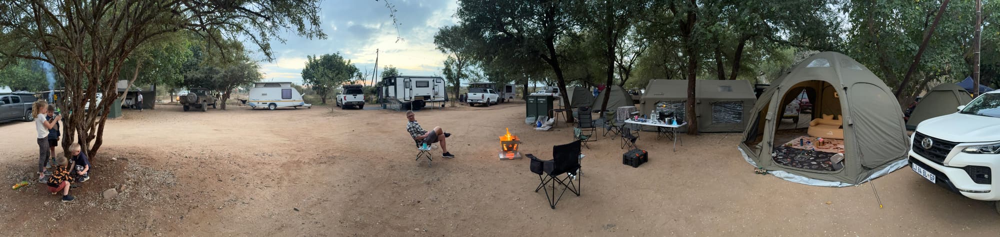 Wide panorama of the campsite at Crocodile Bridge, showing the Coody tents, kids playing, and a fire pit