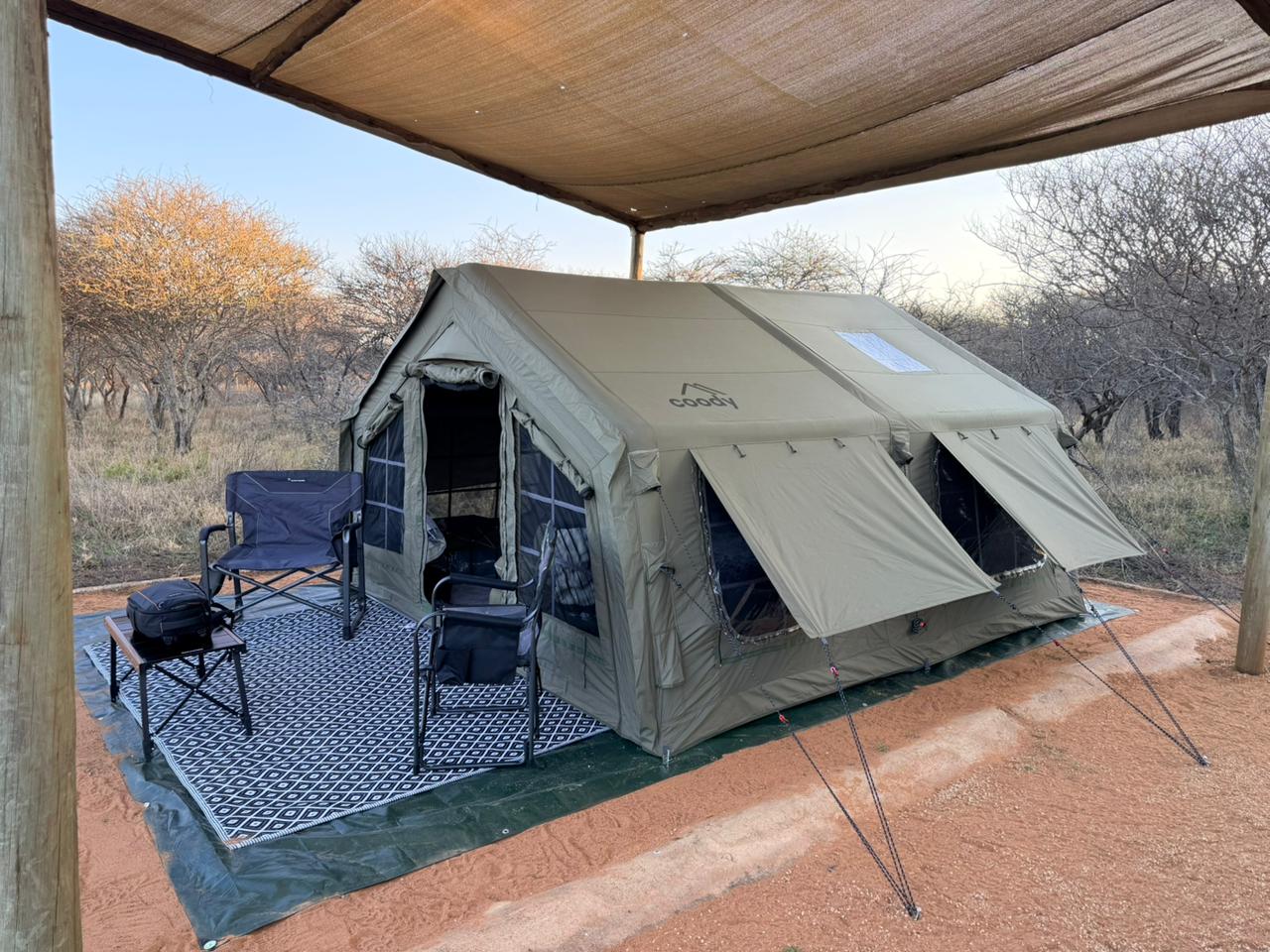 Raudri's army green Coody 13.6 set up under a shade net in the bush, with camp chairs and a rug out front